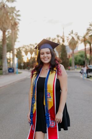 Graduate wearing colorful graduation stole and cap poses on palm tree-lined path during sunset.