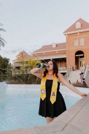 Graduate in academic regalia celebrates by pool with champagne at sunset.