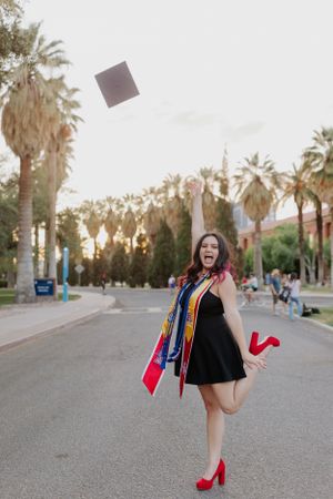 Graduate in black dress and red heels celebrates by throwing cap in air on palm-lined walkway.