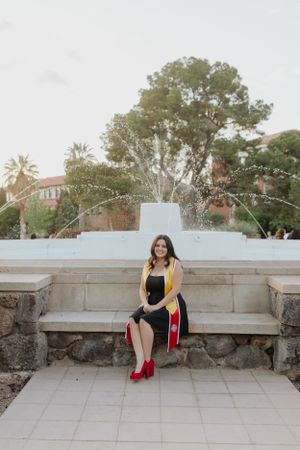 Graduate in academic regalia sitting on stone bench during outdoor photo session.