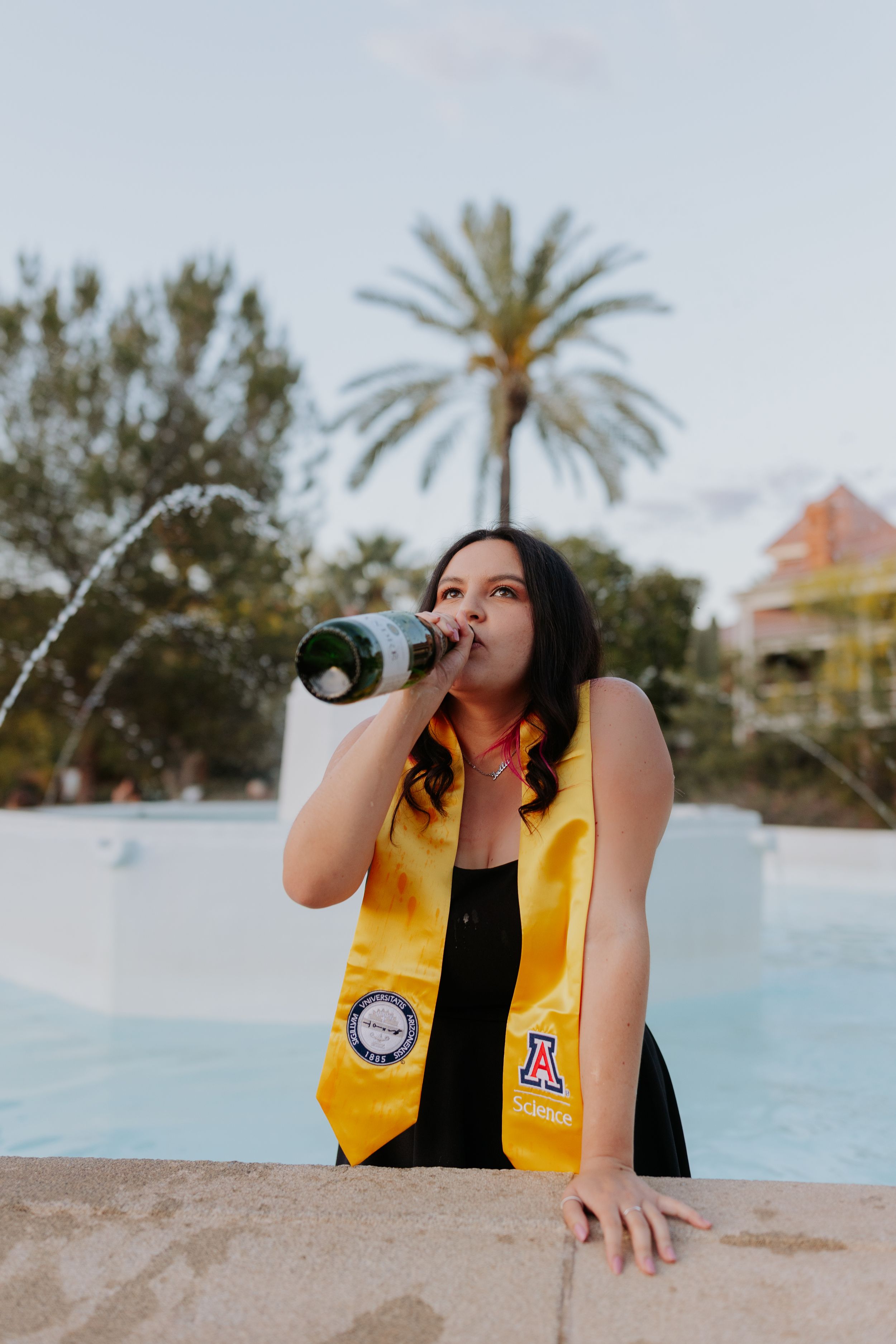 Graduate drinks from champagne bottle during celebratory photoshoot by resort pool.