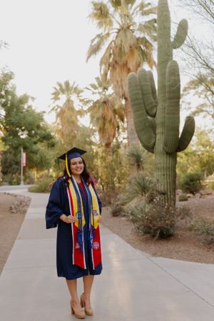 Graduate in academic dress walking along path surrounded by saguaro cacti and palm trees.