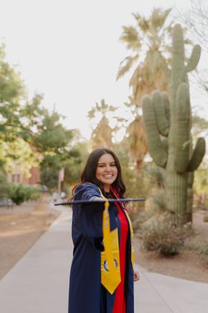 Graduate in academic regalia poses with yellow and red stoles against desert landscape.