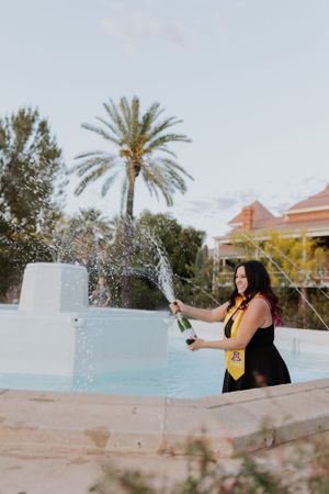 Graduate in black gown and colorful stole sprays champagne by pool with palm trees in background.