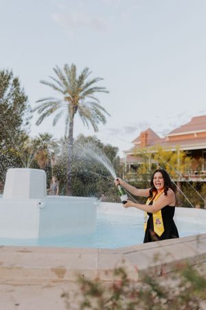 Graduate sprays champagne while celebrating near pool with Spanish-style buildings in background.