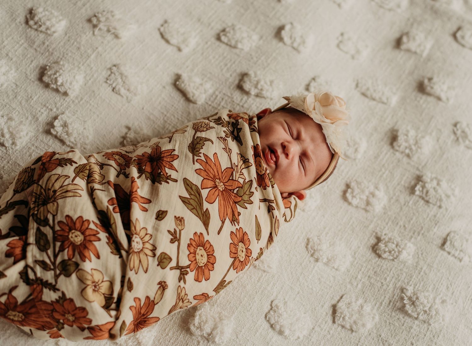 Sleeping newborn swaddled in floral wrap with matching bow headband on textured white blanket.
