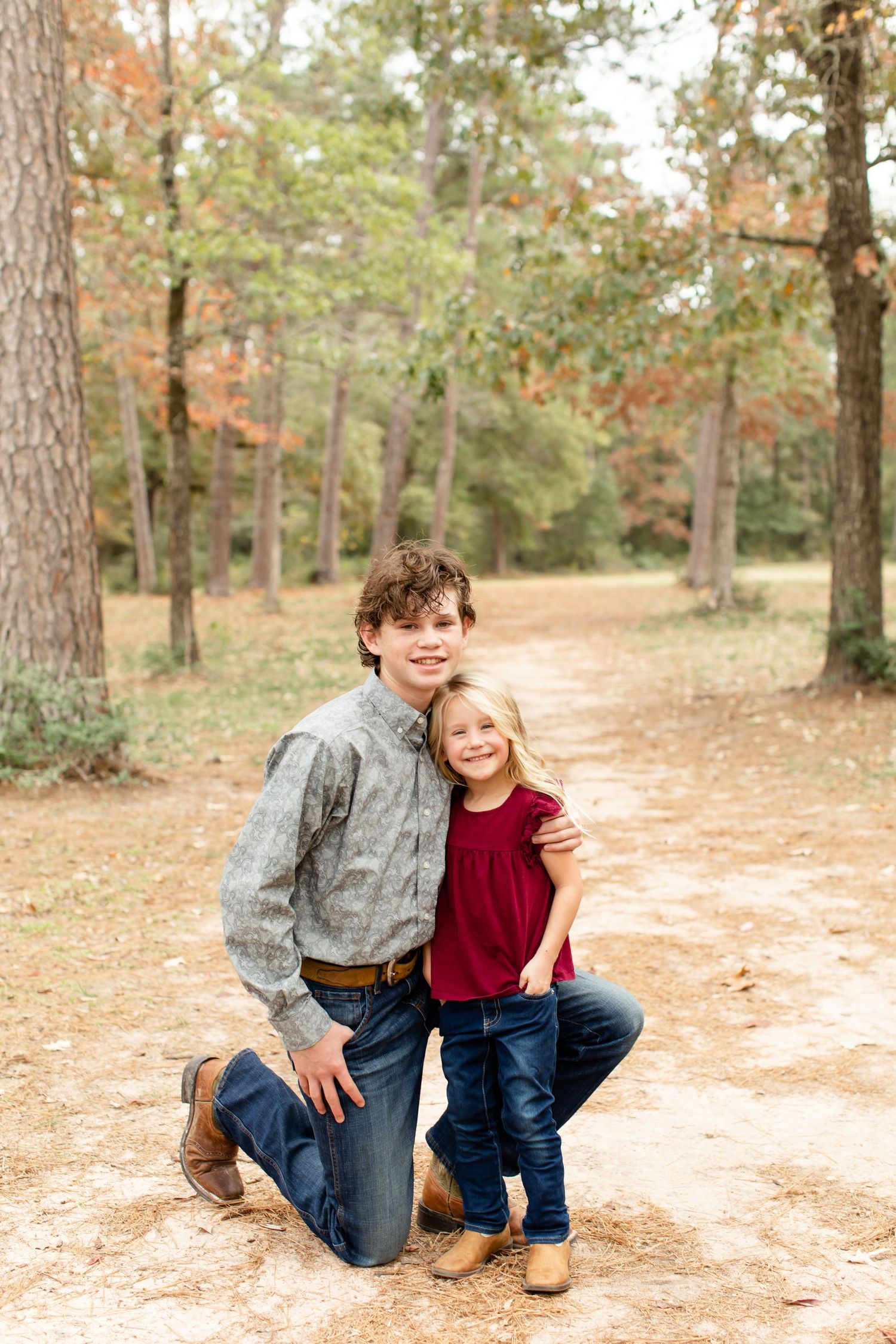 Two siblings pose together on a winding dirt path in an autumn forest setting.