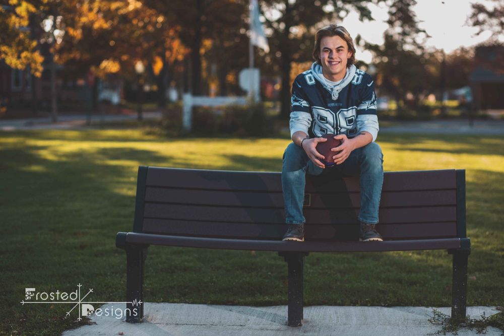 High School Senior boy holding football on park bench with a football jersey on.