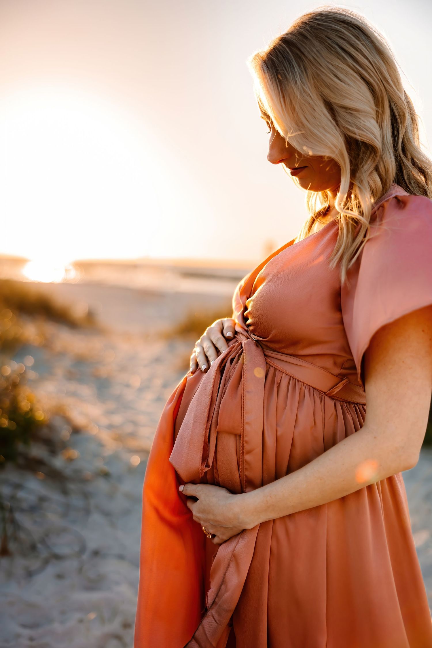 Pregnant woman in a terracotta dress cradling her baby bump at a sandy beach during sunset.