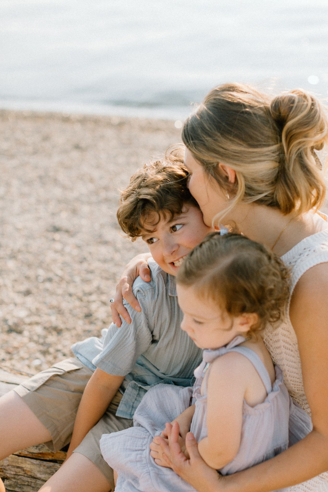 A mother sits on a pebble beach embracing her two young children, sharing a tender moment in soft golden light.