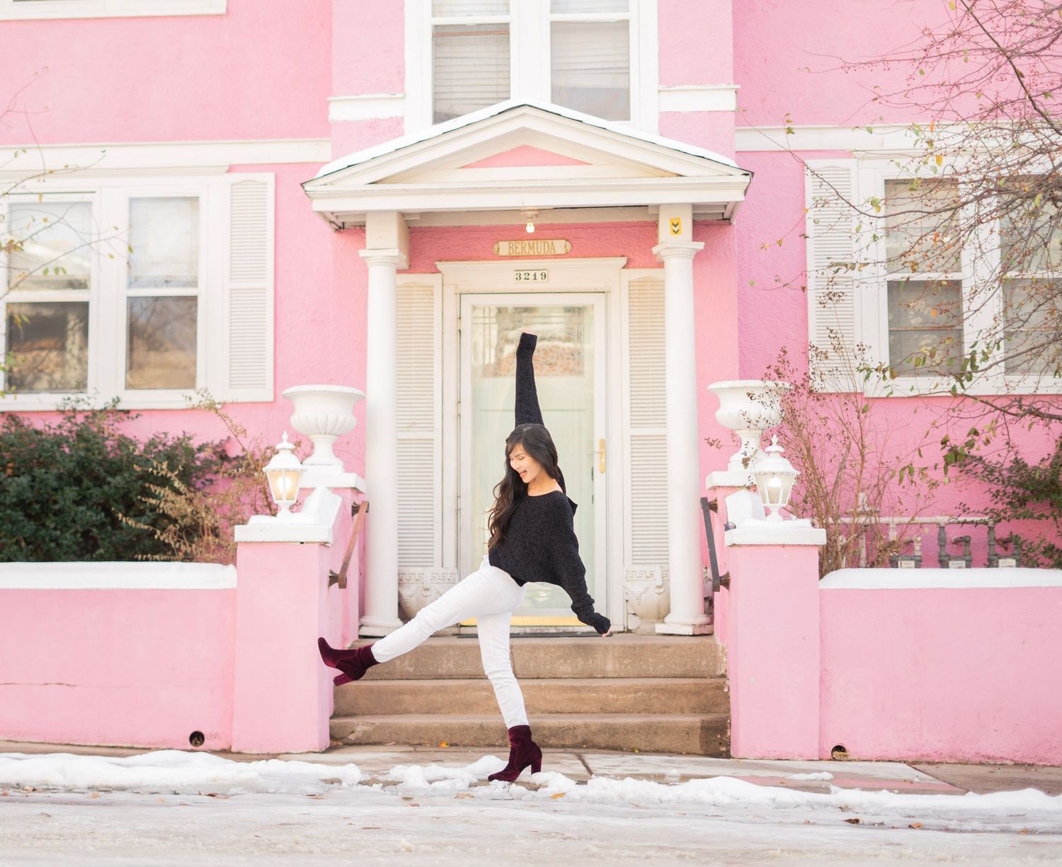 A dancer poses gracefully in front of a pink building in winter, wearing a black sweater, white pants, and burgundy boots.