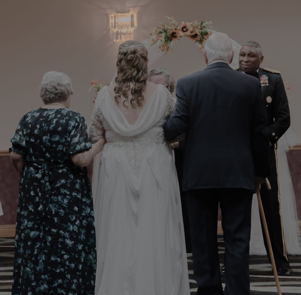 A wedding ceremony viewed from behind showing attendees facing a decorated arch with floral arrangements.