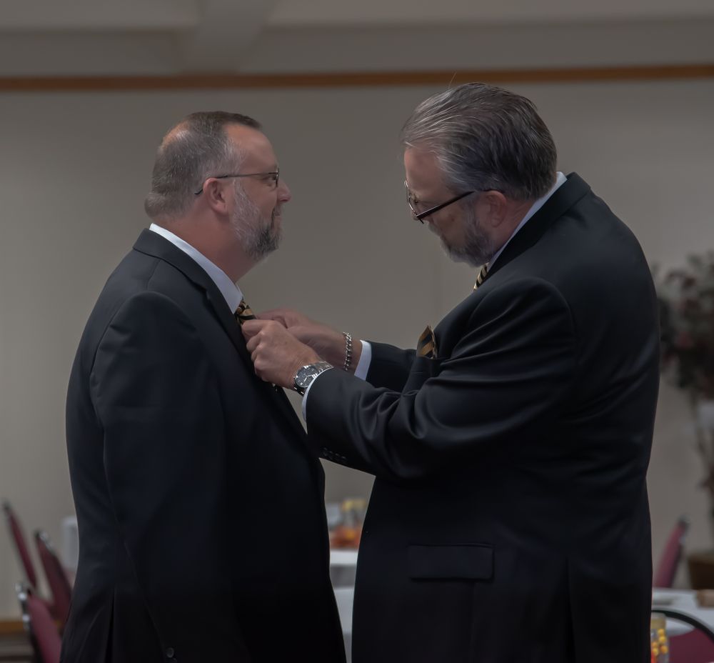 Two men in suits adjust formal attire at an indoor event.