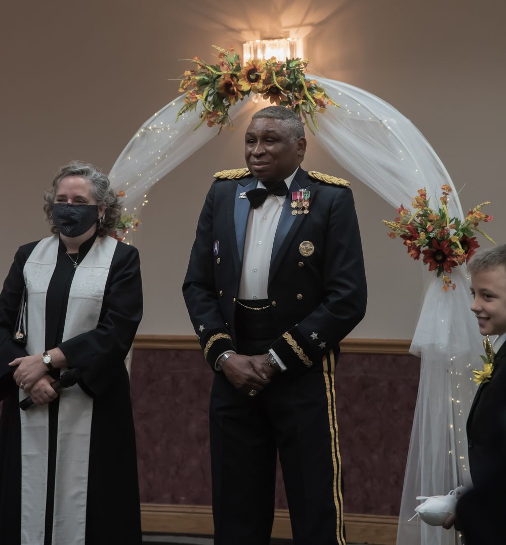A military officer in dress uniform stands at a wedding ceremony under a decorated arch with floral arrangements.