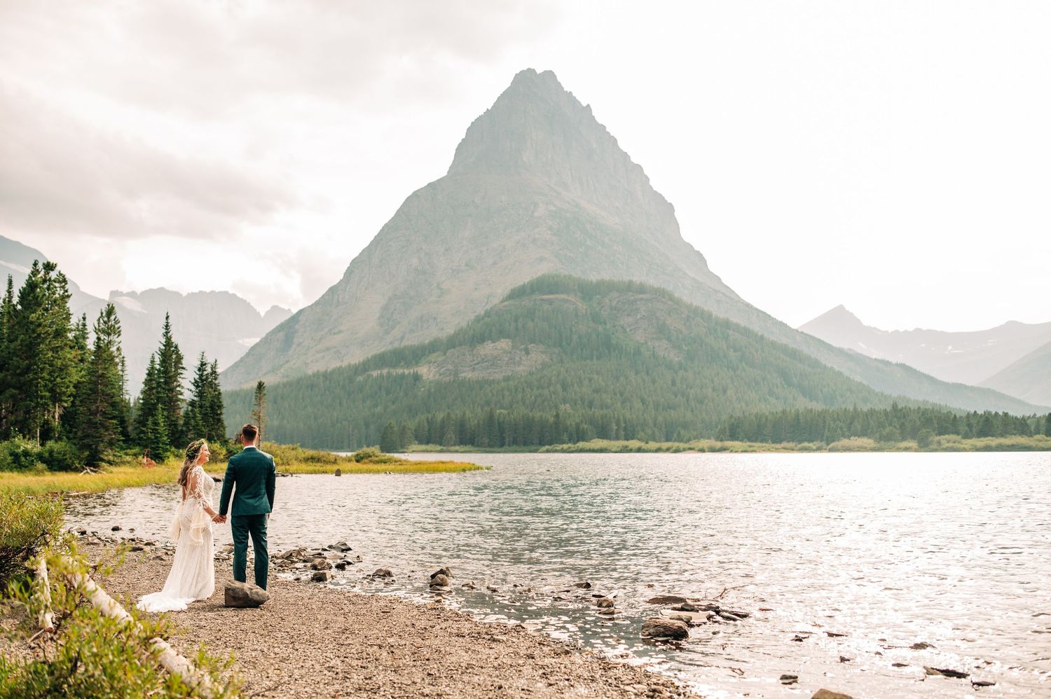 A couple walks along a lakeside trail with iconic Mount Grinnell towering in the background.