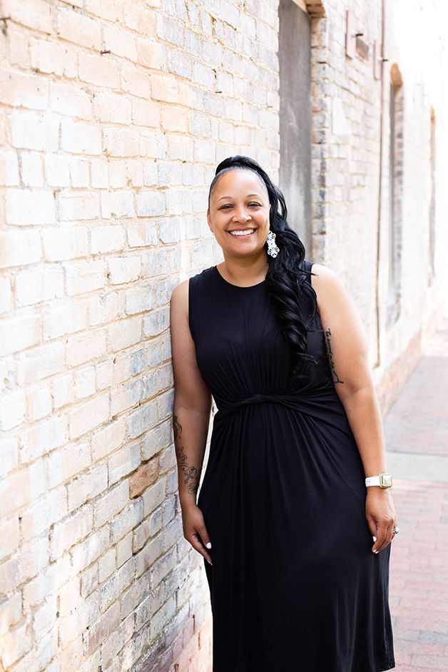 Woman in black dress leaning against a white brick wall smiling at the camera.