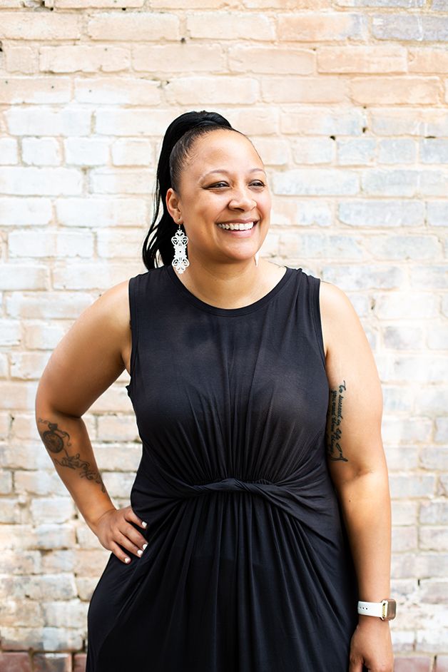 Woman wearing a black dress with white brick in the background.  She has hand on her hip looking slightly off camera.