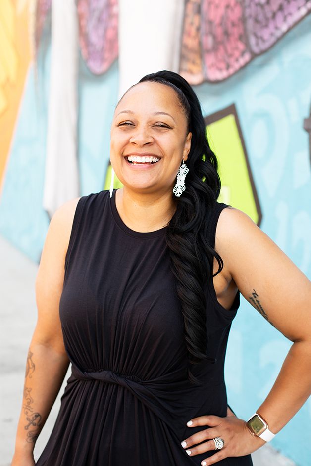 Woman is black dress headshot with colorful mural in background laughing at the camera.
