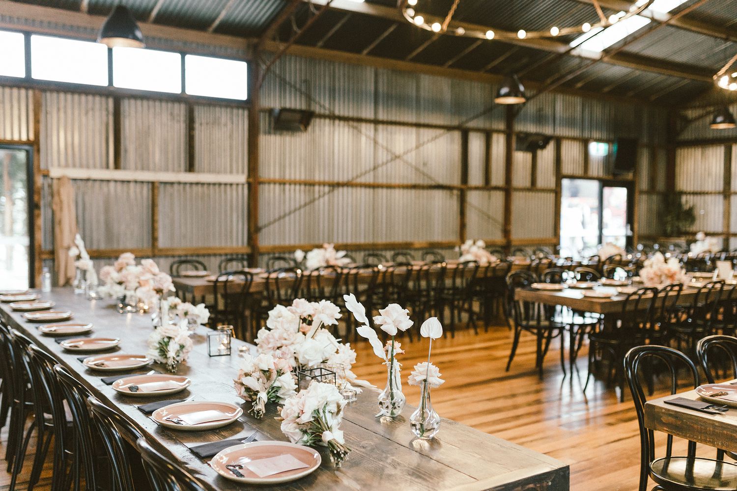 Wedding reception setup in industrial-style venue with long wooden tables and black chairs.