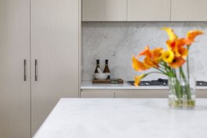 Minimalist kitchen with white cabinets, marble backsplash, and orange lilies in vase.