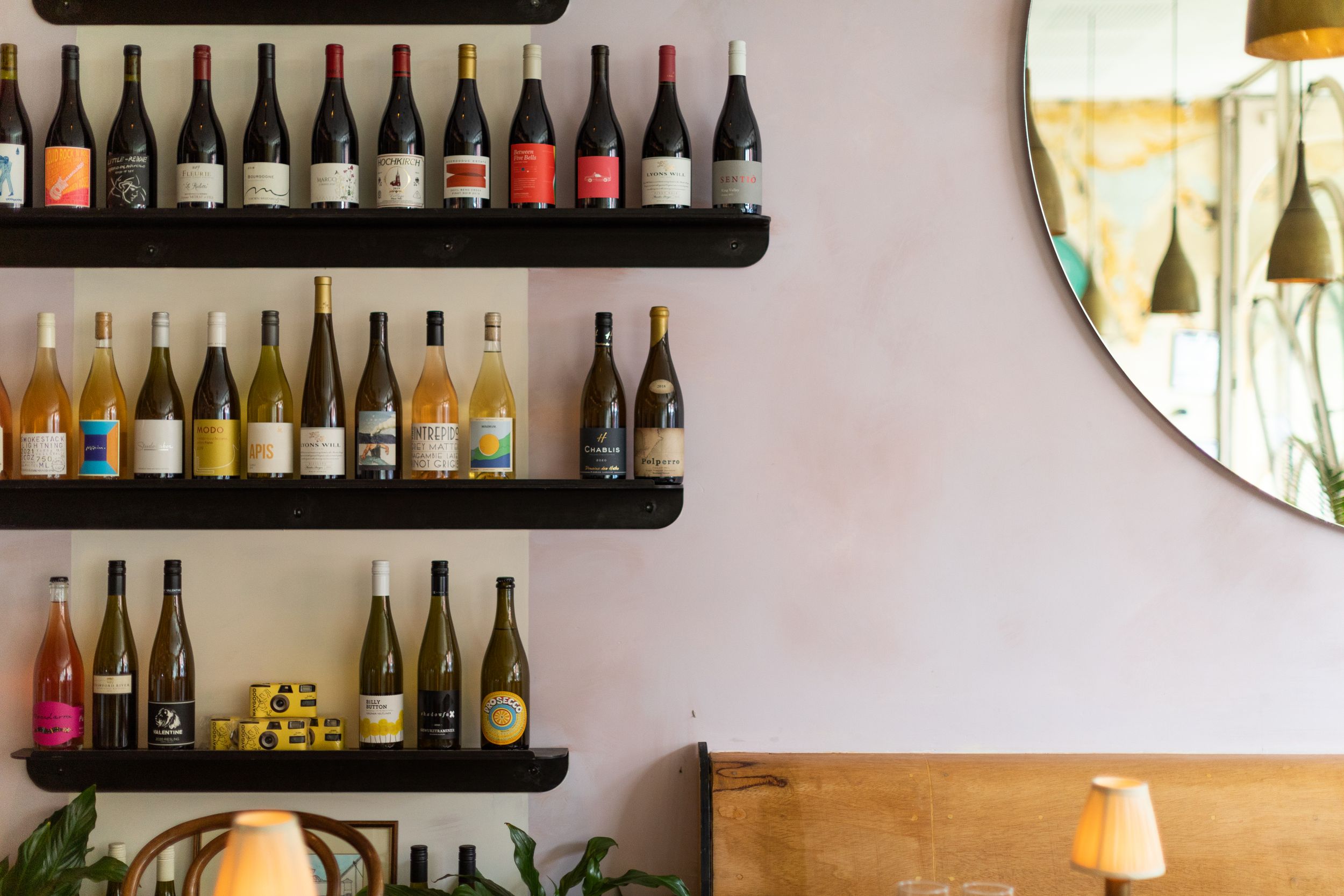Wine bottle display on black floating shelves against pink wall with round mirror.
