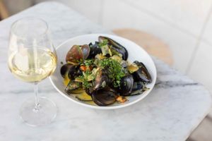 Plate of steamed mussels with vegetables served alongside white wine on marble table.