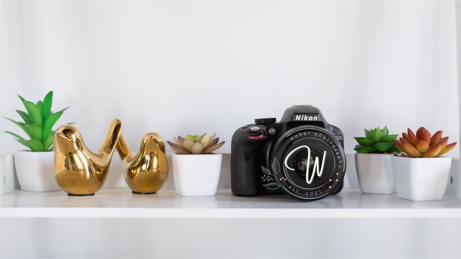 Modern white shelf displaying a DSLR camera, small succulents in white pots, and gold decorative objects.