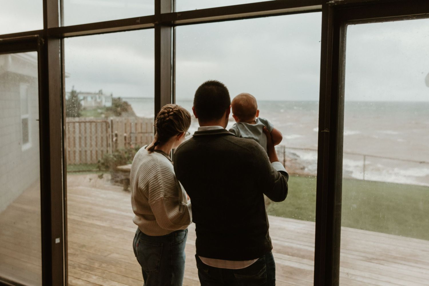 Couple admiring coastal view through large window while holding baby on overcast day.