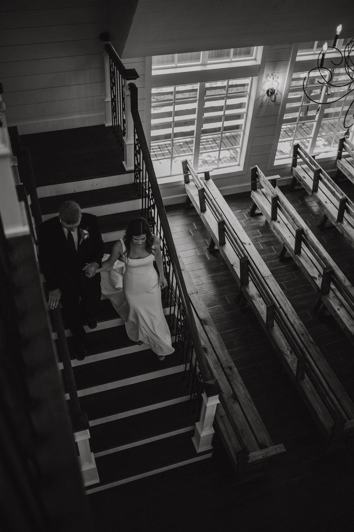 Black and white photo of couple walking down church stairs with wooden pews visible.
