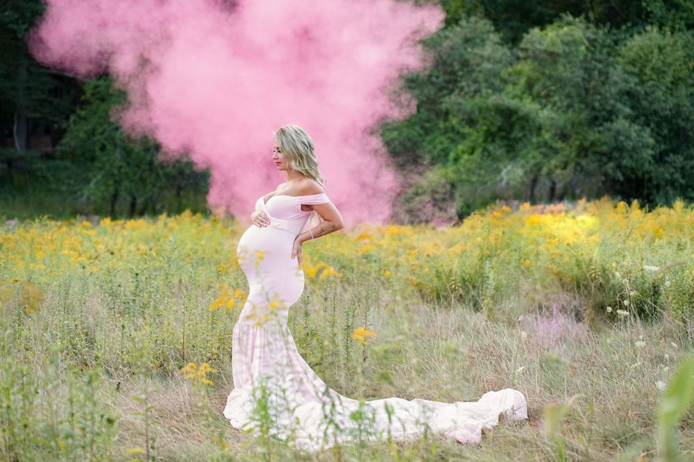 Creative maternity photo with pink smoke effect in field of yellow wildflowers.