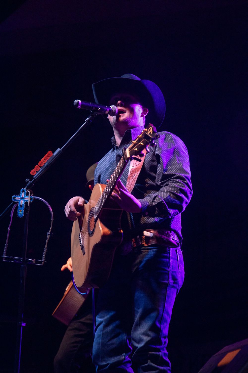 Country musician in cowboy hat singing and playing acoustic guitar under purple stage lighting.