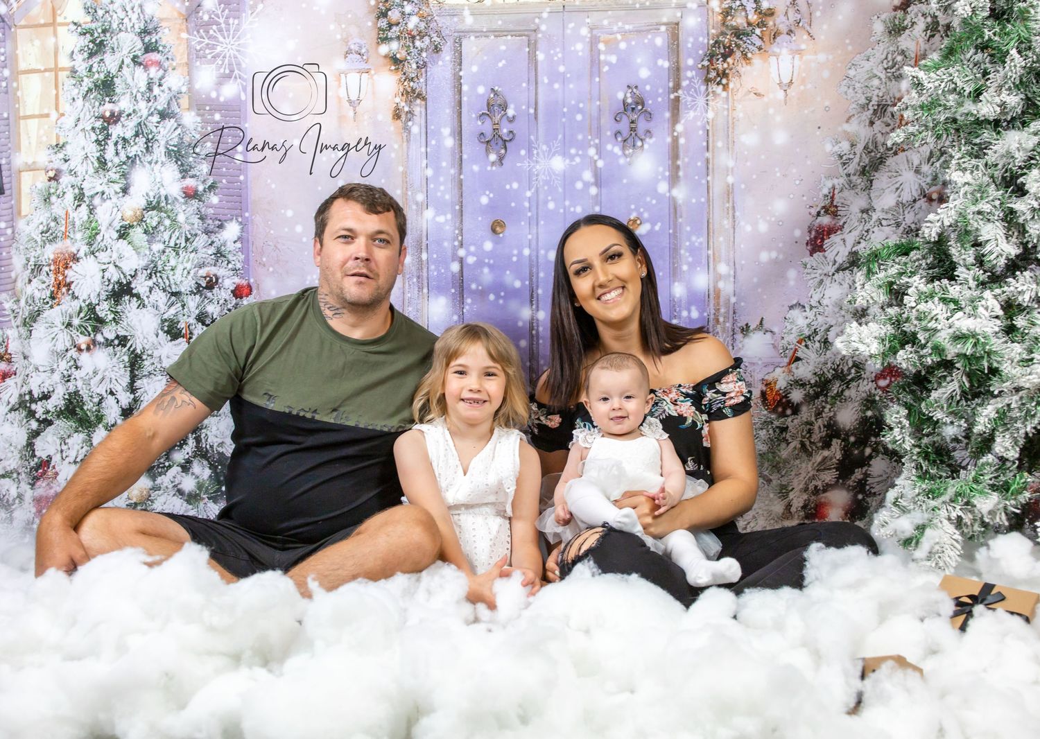 Family of four poses in a festive Christmas photo session with decorated trees and snow.