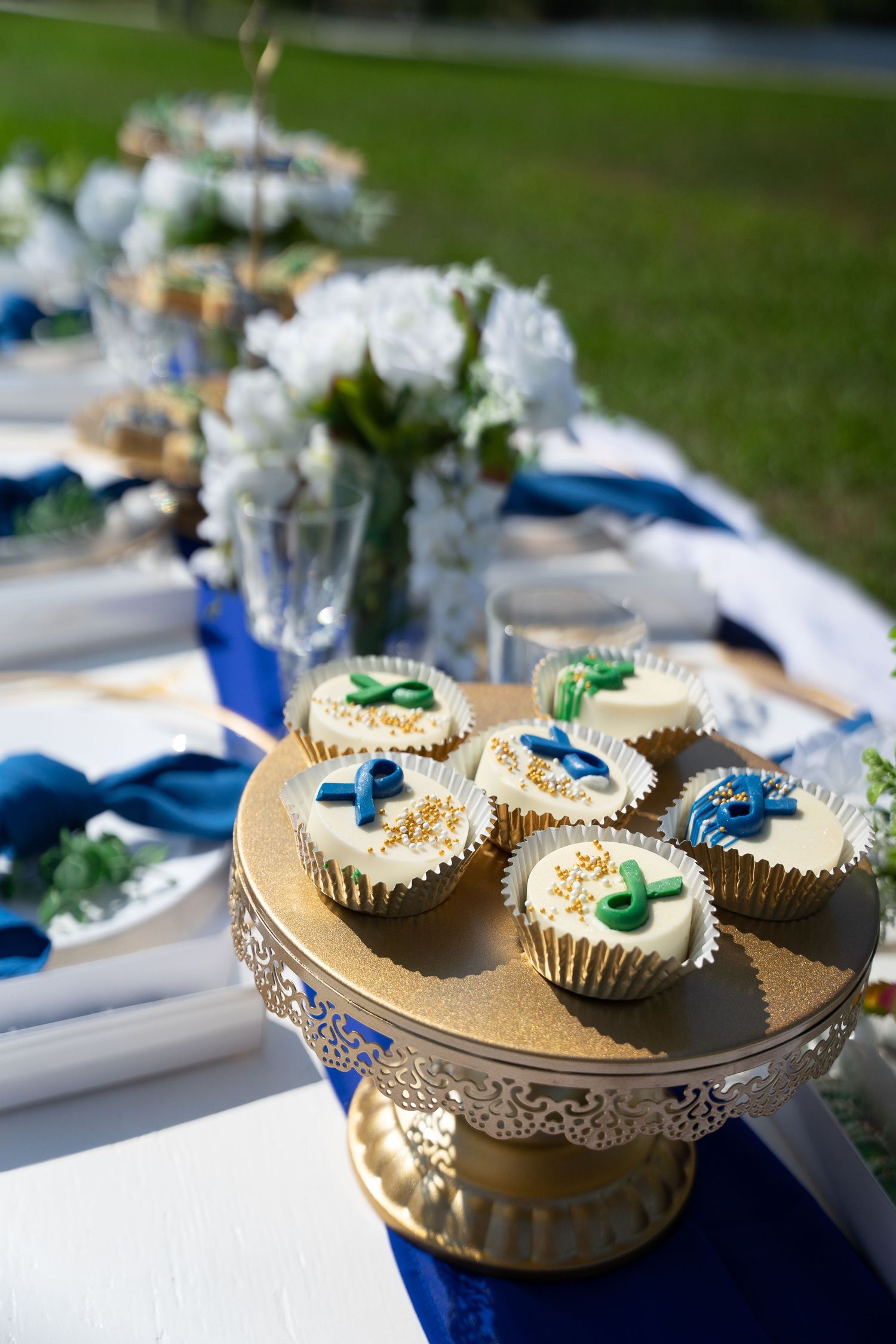 Elegant cupcakes with blue and green decorations displayed on a gold cake stand at an outdoor event.