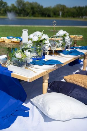 Lakeside outdoor dining setup with blue and white decor, pillows, and floral arrangements.