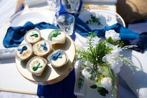 Decorated cupcakes on a gold stand next to white floral arrangements with blue accents.