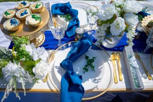 Formal place setting with gold flatware, blue ribbons, and white floral centerpieces.
