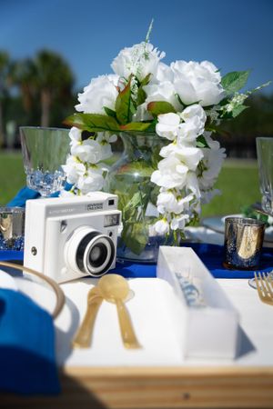 Elegant white flowers and vintage camera decorate a blue and white tablescape at an outdoor event.