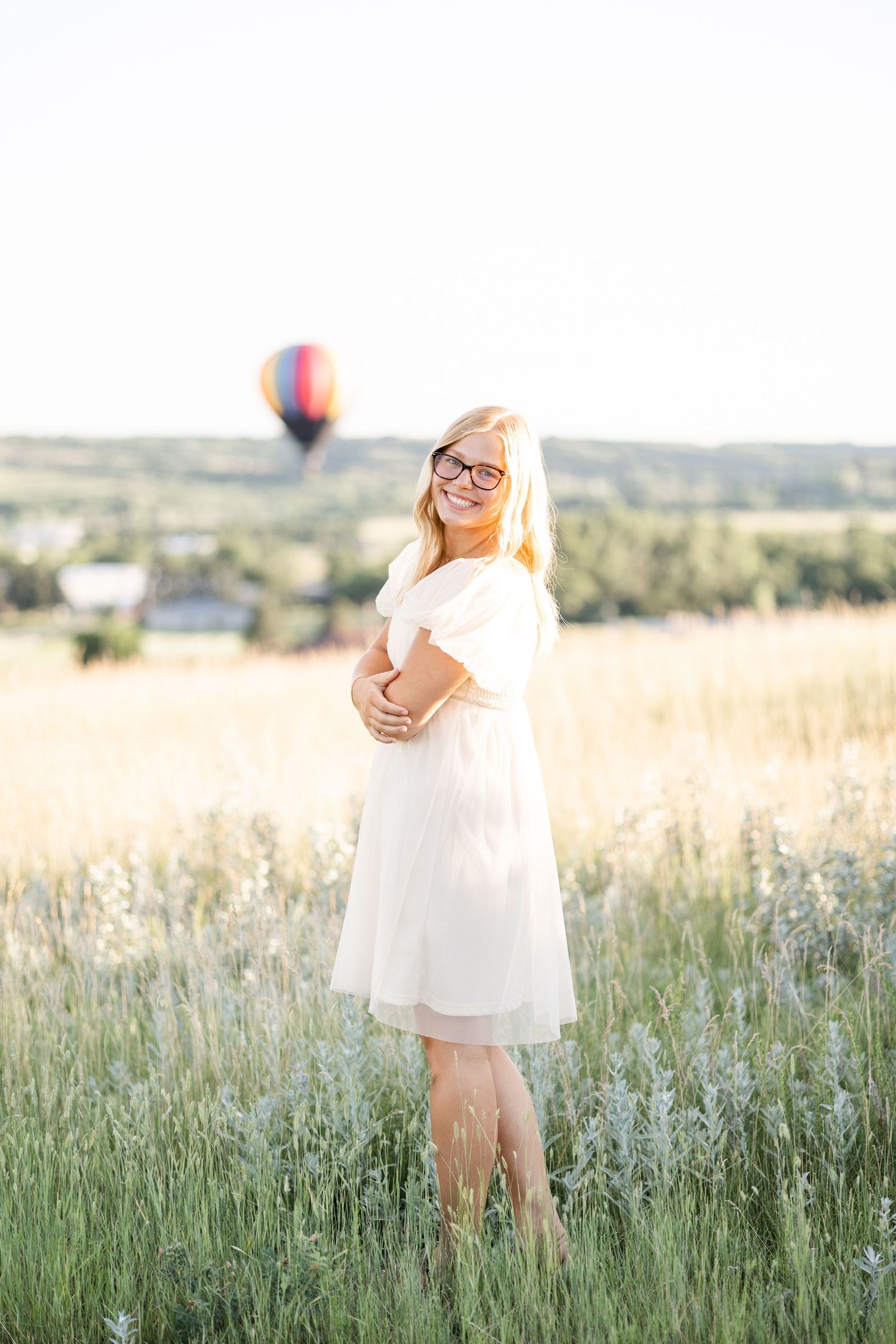 Woman in white dress stands in meadow with hot air balloon floating in distance.