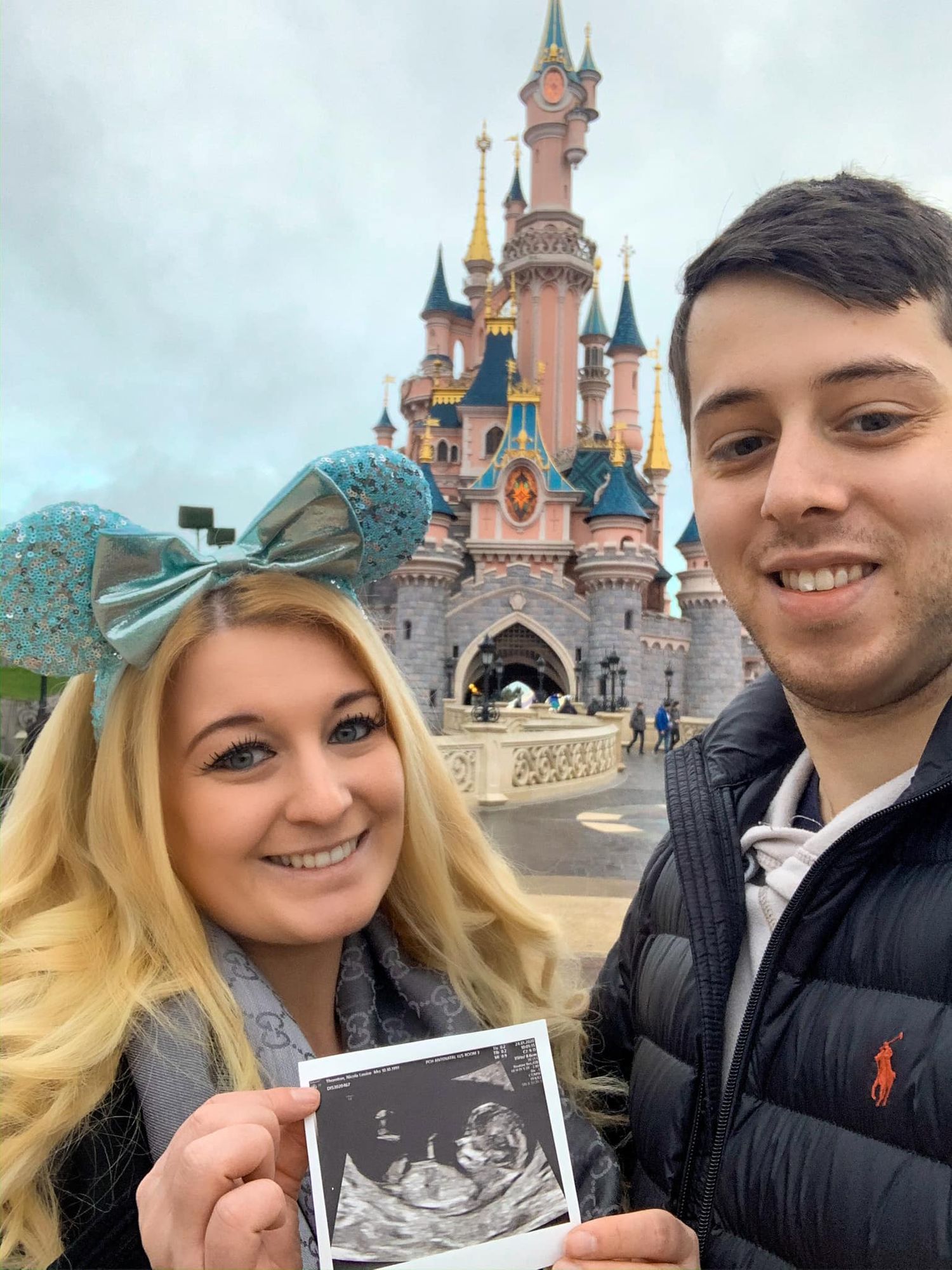 A couple poses with an ultrasound photo in front of the iconic Disneyland castle.
