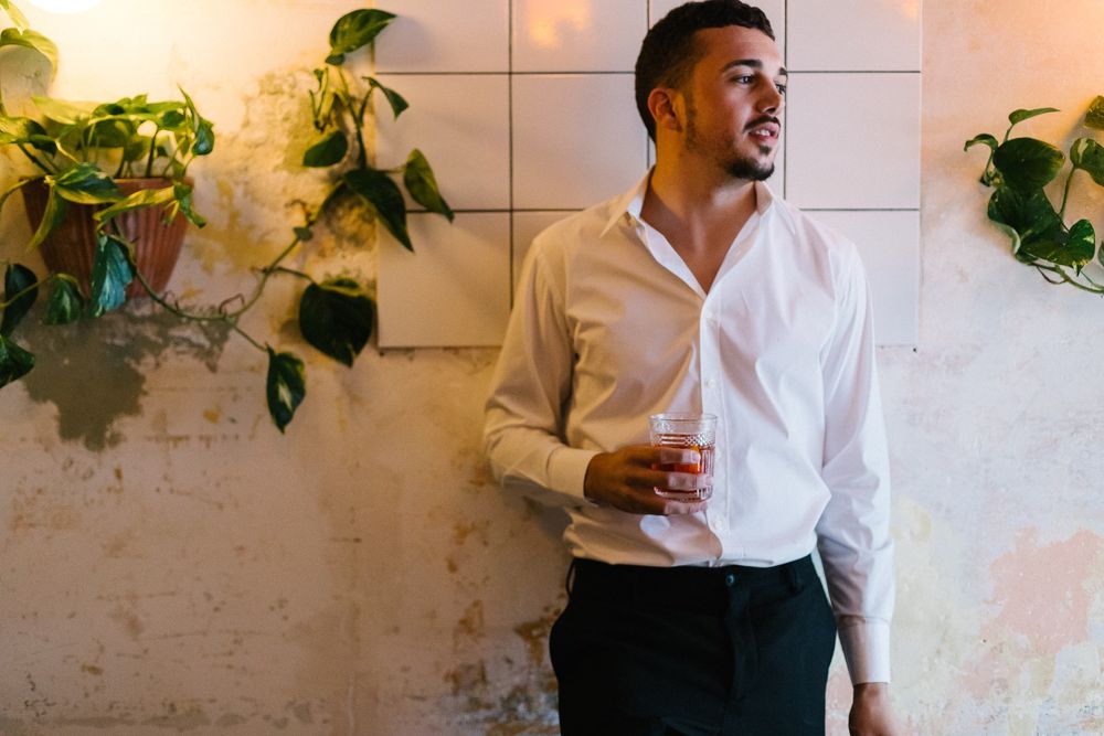 Fashionable person in white shirt and dark pants standing against textured wall with green ivy plants.