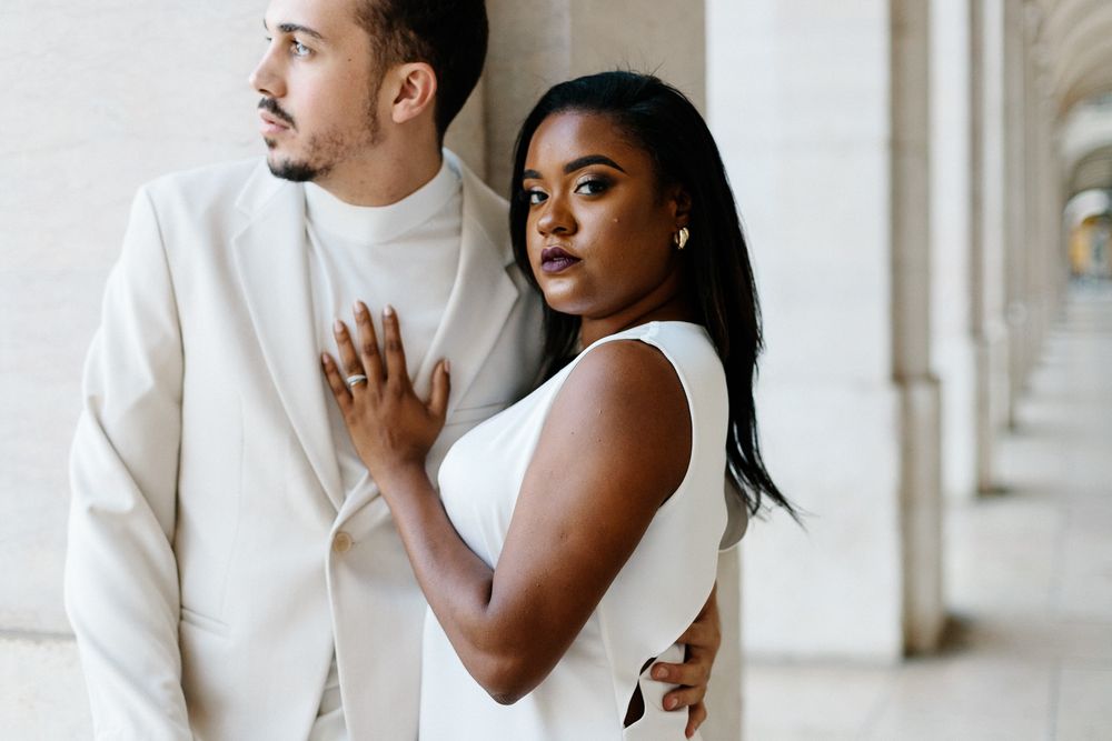 Stylish couple in white outfits embrace while posing in front of architectural columns.