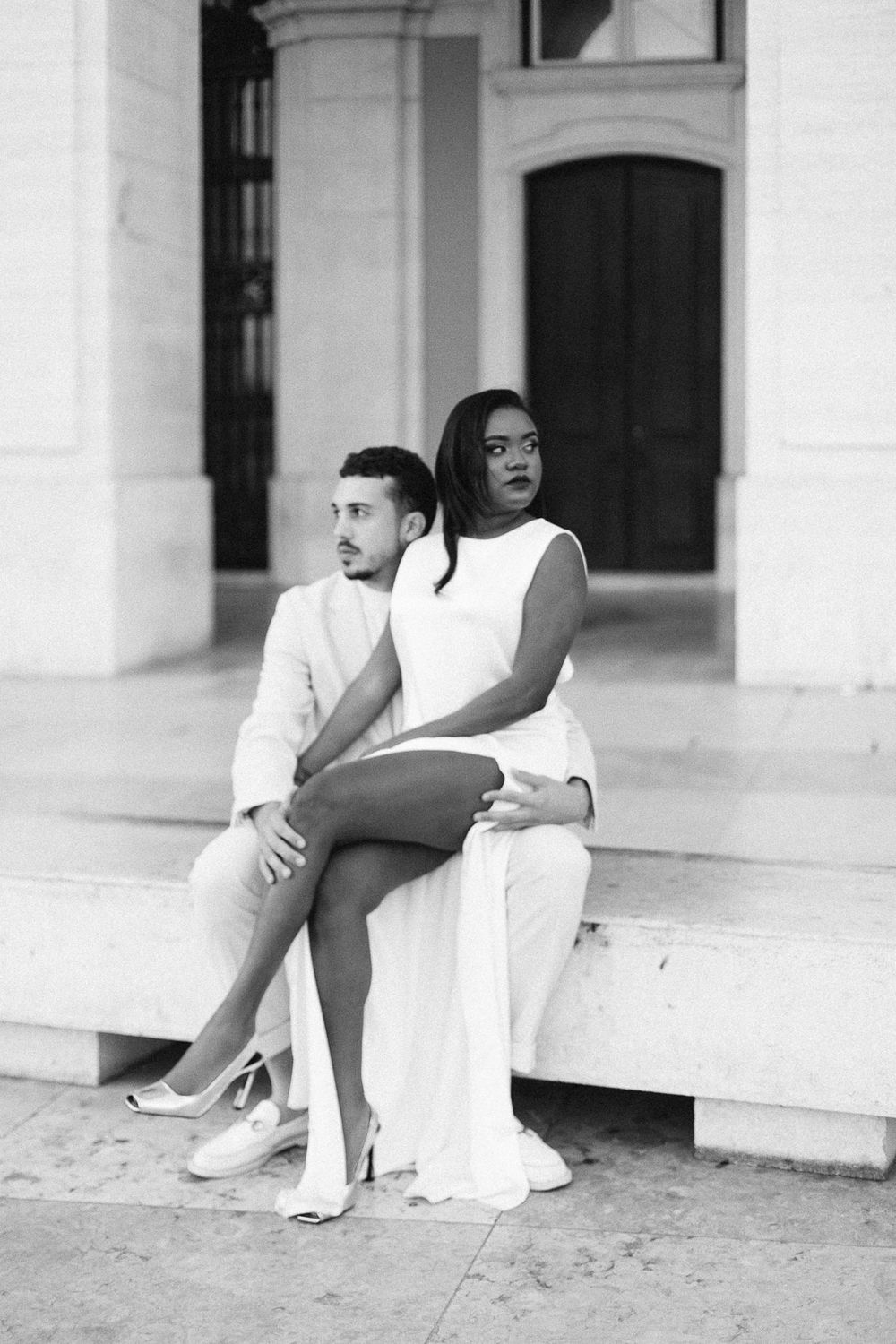 Stylish couple in white outfits poses on marble steps of historic building.