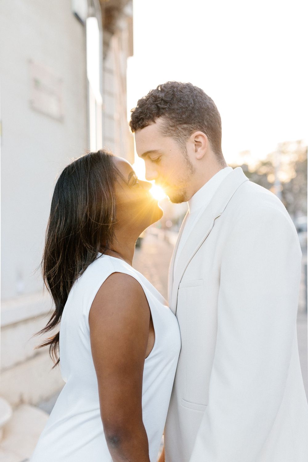 Intimate couple in white outfits share a tender moment at sunset with golden light streaming between them.