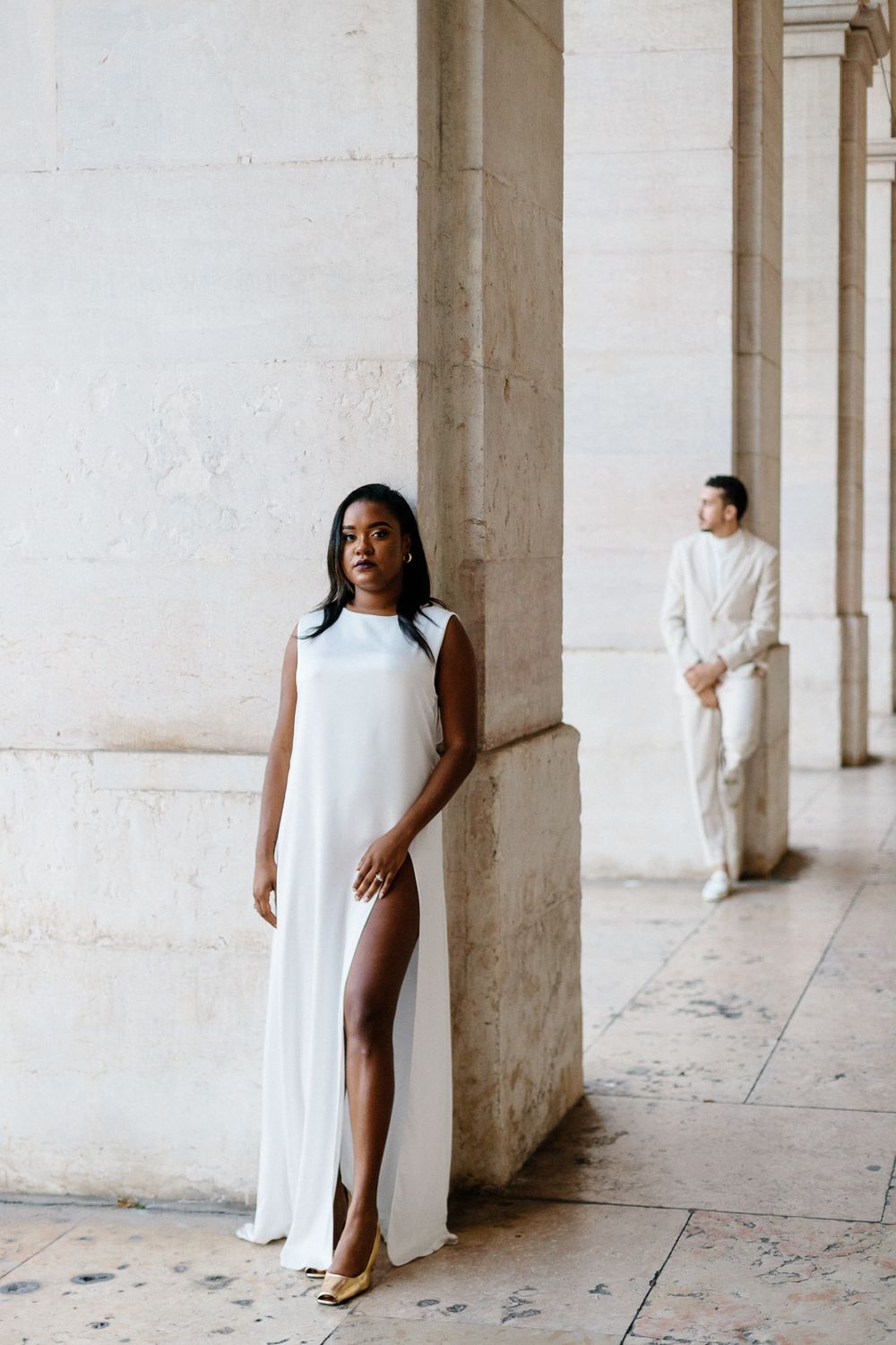 Couple in white formal wear pose separately in elegant colonnade setting.