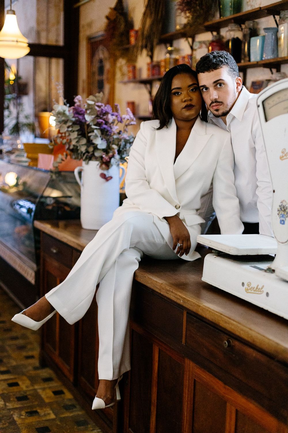Elegant couple in white outfits posing at vintage counter with floral arrangements.