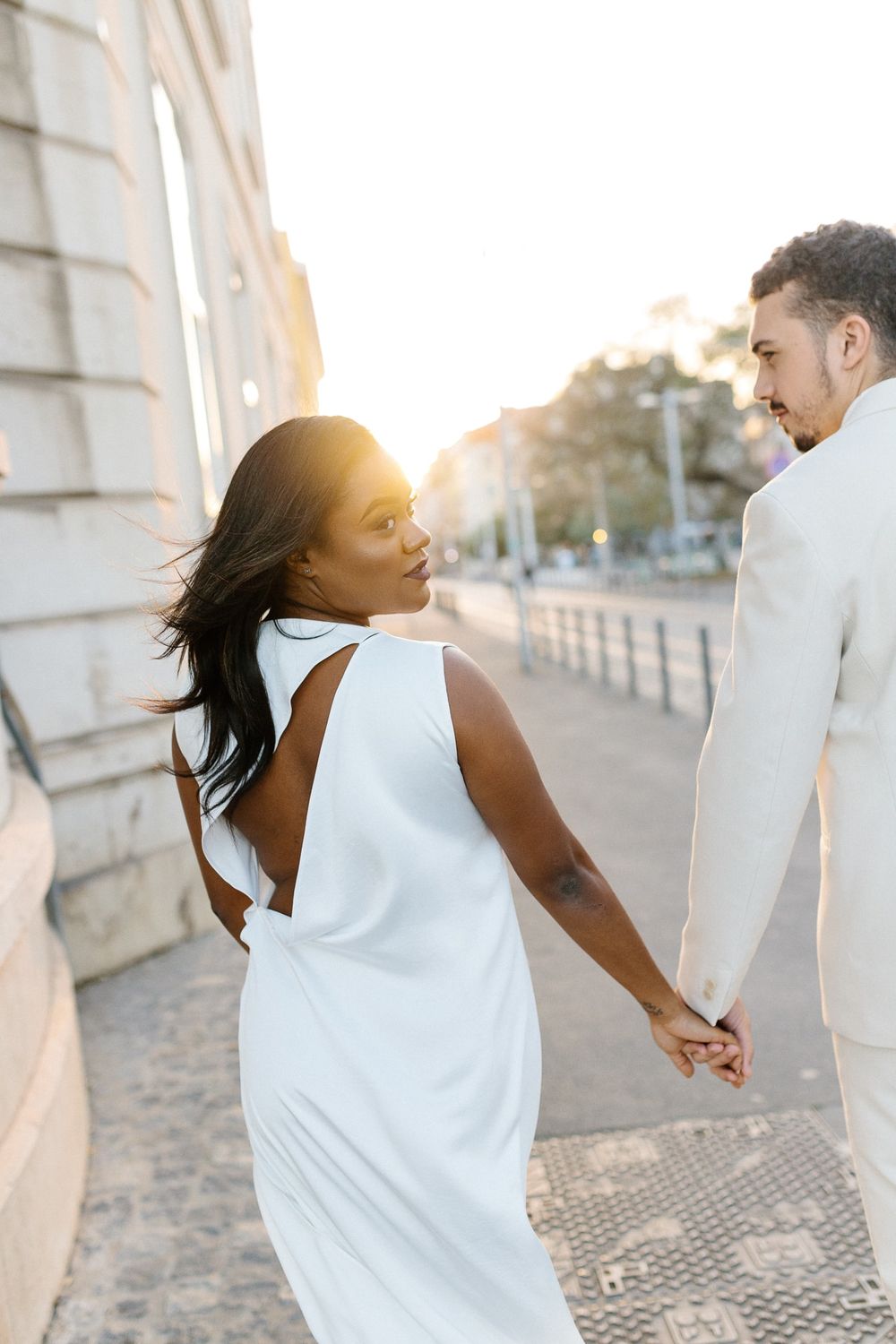 Couple in coordinating white outfits holding hands while walking along a sunlit path.