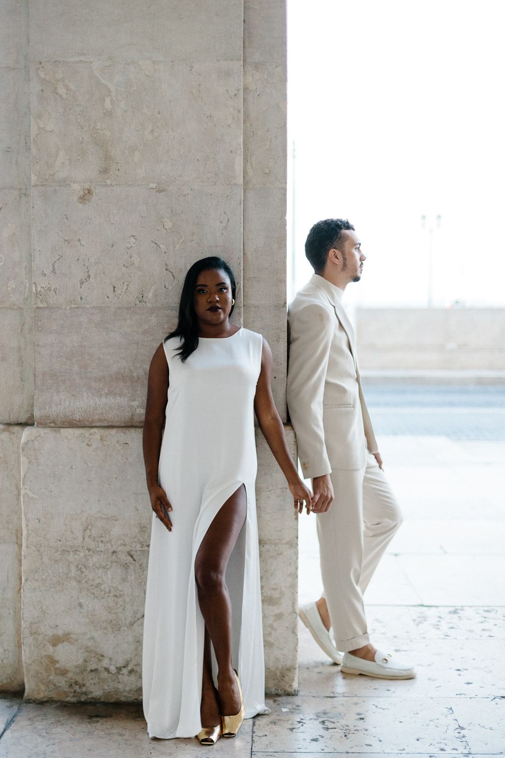 Couple in white holding hands while posing against stone column backdrop.