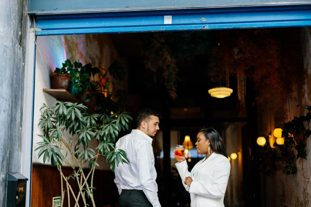 Person in white blazer enjoying red cocktail in dimly lit venue with plants.