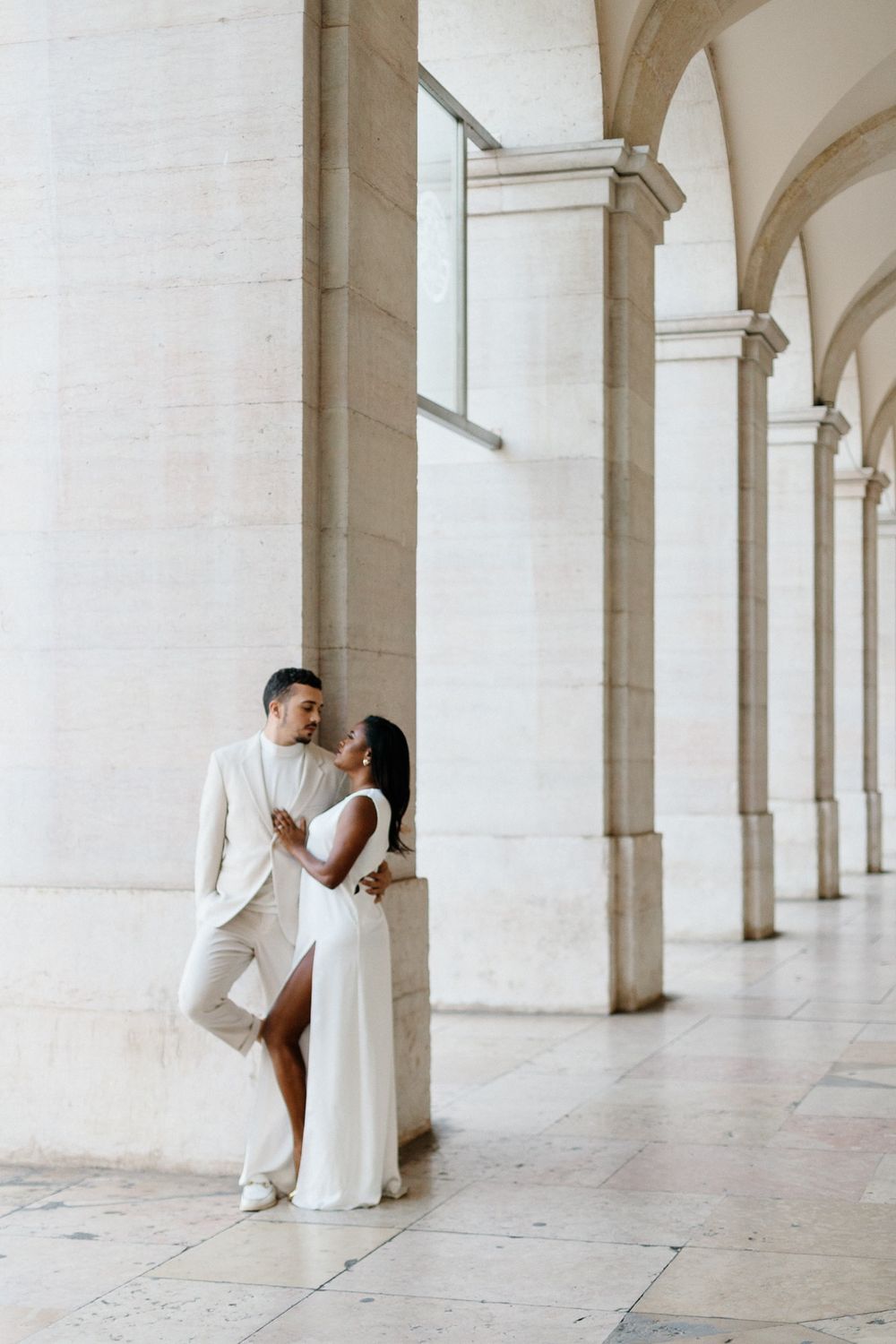 Couple in elegant white attire pose romantically against classical stone columns.