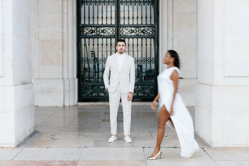 Fashionable couple in white outfits pose playfully in front of ornate iron gate.